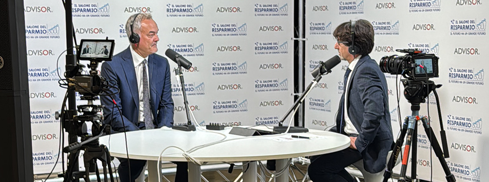 Intervista in regia durante un evento: due ospiti in cuffia al tavolo con camere su treppiede, microfoni a braccio e backdrop brandizzato