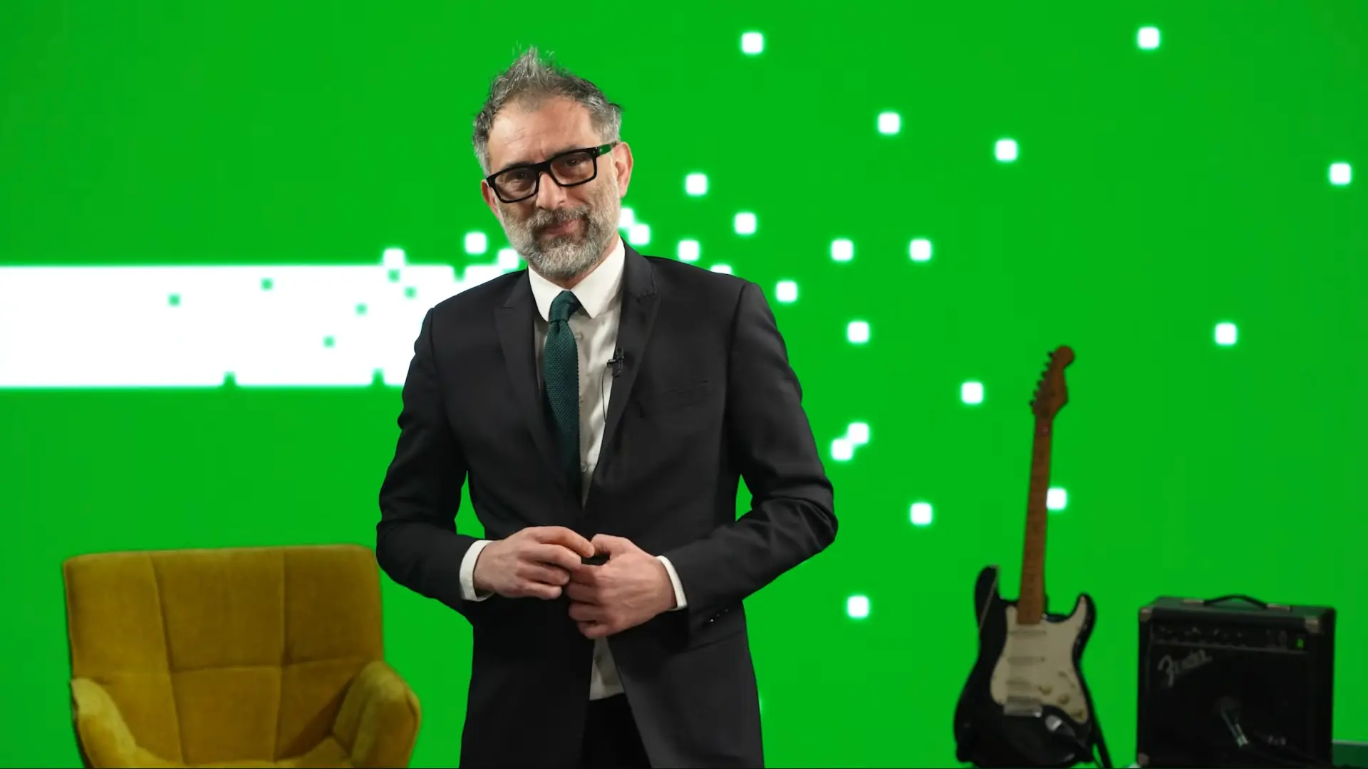 Uomo in completo scuro davanti a una parete LED verde con grafiche a pixel bianchi, poltrona in velluto e chitarra in studio