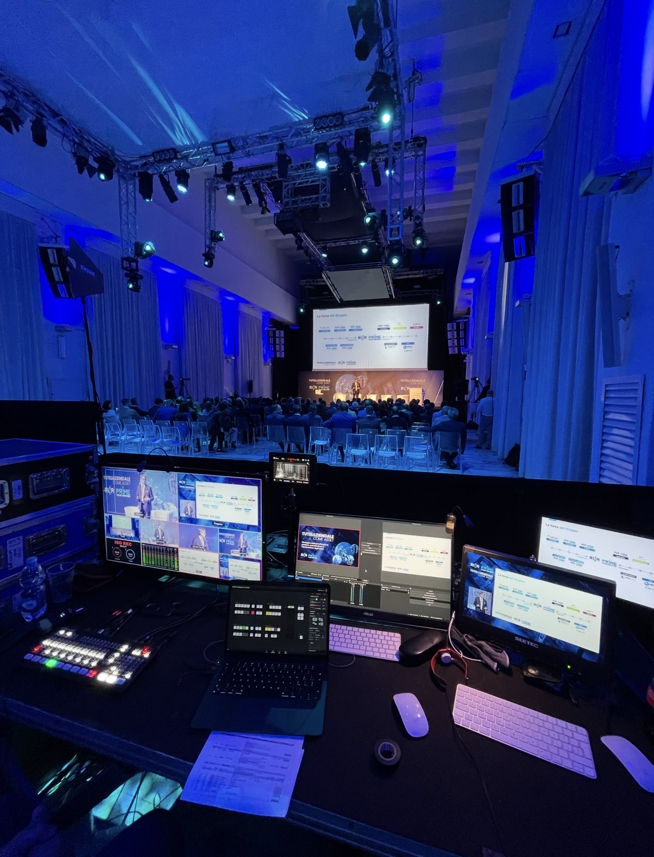 Regia multicamera live da una cabina di controllo durante un evento aziendale: monitor, mixer video e palco illuminato in blu