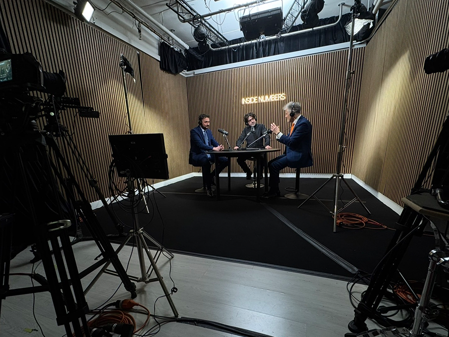 Camere sul set: tre ospiti al tavolo ripresi da una camera professionale su treppiede, con softbox e pannello LED in regia
